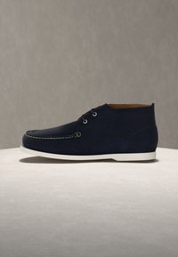 Navy blue suede lace-up men's ankle shoe with white sole and contrast stitching on a neutral background.