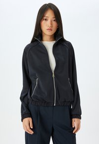 Svart zip-jacka med hög krage, elastisk midja och sidofickor. Har en slät yta och subtil glans.