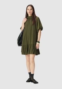Vestido verde oliva, a la altura de la rodilla, con un cuello con volantes y mangas cortas abullonadas. La modelo lleva un bolso negro, usando calcetas negras con lunares y zapatos planos.