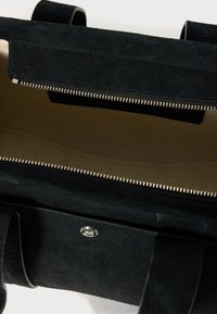 Schwarze Wildleder-Tasche mit einem Reißverschluss, beigem Innenfutter und einem silbernen Logo-Akzent auf der Vorderseite. Minimalistisches Design, rechteckige Form.
