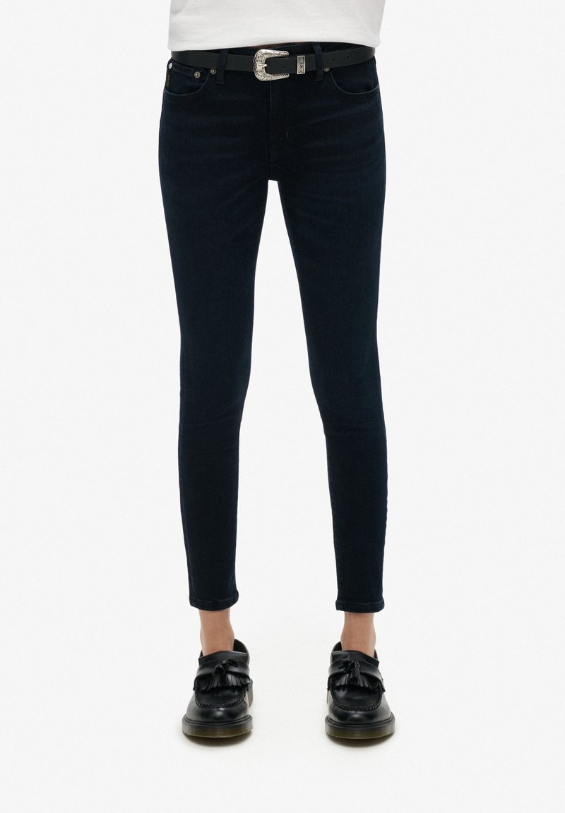 Jeans skinny bleu foncé avec une texture lisse, cinq poches, et une ceinture à boucle argentée, associés à des chaussures noires à enfiler.