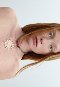 Collana con pendente a forma di stella in metallo con superficie testurizzata, cordoncino in tessuto verde e perline viola, caratterizzata da un design asimmetrico e tonalità naturali.