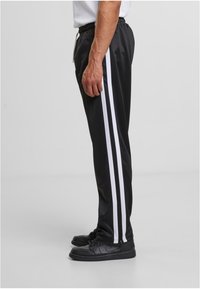 Crne trenirka napravljene od glatkog materijala, s dvije vertikalne bijele pruge s obje strane, s elastičnom trakom u struku i patentnim zatvaračima na gležnjevima.