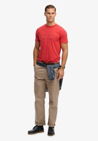 Czerwona bawełniana koszulka z czarnym nadrukiem, beżowe spodnie, niebieska kraciasta koszula zawiązana wokół talii oraz czarne sznurowane buty z teksturowaną podeszwą.