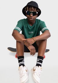 Grünes sportliches T-Shirt mit weißen Streifen, schwarze Shorts, schwarze und weiße Nike-Socken, Sneakers mit goldenen Akzenten, schwarzer Bucket Hat und große Sonnenbrille.
