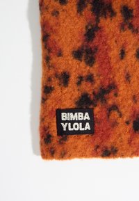 Tela de fieltro en naranja y rojo con manchas negras. Presenta una etiqueta con texto blanco sobre un fondo negro: "BIMBA YLOLA."