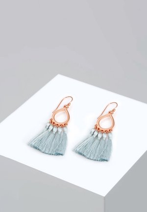 Boucles d'oreilles en forme de goutte en or rose avec des pampilles en tissu bleu clair, présentées sur un cube blanc sur un fond gris.