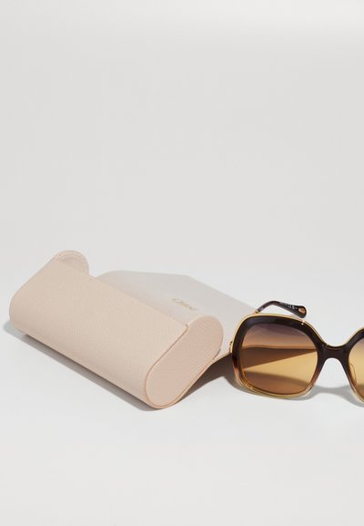 Gafas de sol con lentes marrones degradados y grueso marco de carey al lado de un estuche de cuero texturizado beige con bordes redondeados y un logo dorado.
