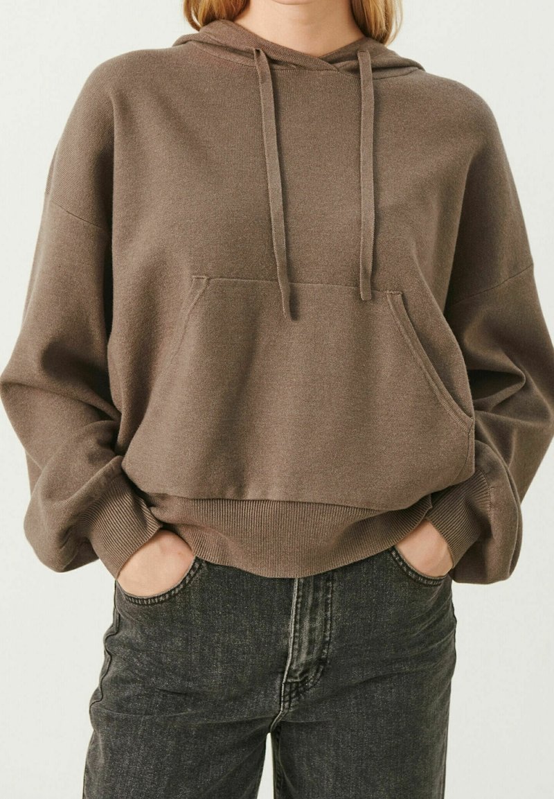 Brown hooded sweatshirt with a front pocket, ribbed cuffs, and hem. Worn with dark jeans, featuring a relaxed fit and soft texture.