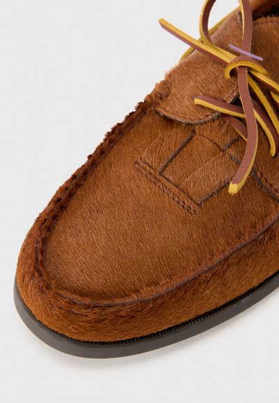 Scarpa in suede marrone con una finitura morbida e strutturata, caratterizzata da un design con lacci e dettagli di cucitura visibili lungo la suola.
