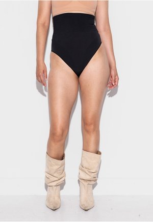 Moterys dėvi aukštakulnius juodus apatinius, smėlio spalvos trumpą palaidinę ir laisvus smėlio spalvos zomšinius batus, stovinčią priešais baltą foną.