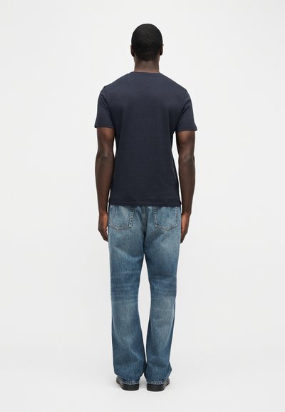 Άνδρας που στέκεται με την πλάτη στην κάμερα, φοράει ένα σκούρο μπλε μπλουζάκι και ανοιχτό μπλε τζιν με ίσια γραμμή, σε απλό λευκό φόντο.