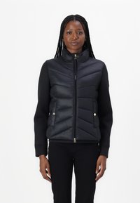 Vest puffer negru cu quilting în zigzag, guler înalt și închidere cu fermoar. Mâneci lungi dintr-un material neted. Accente de hardware aurie.