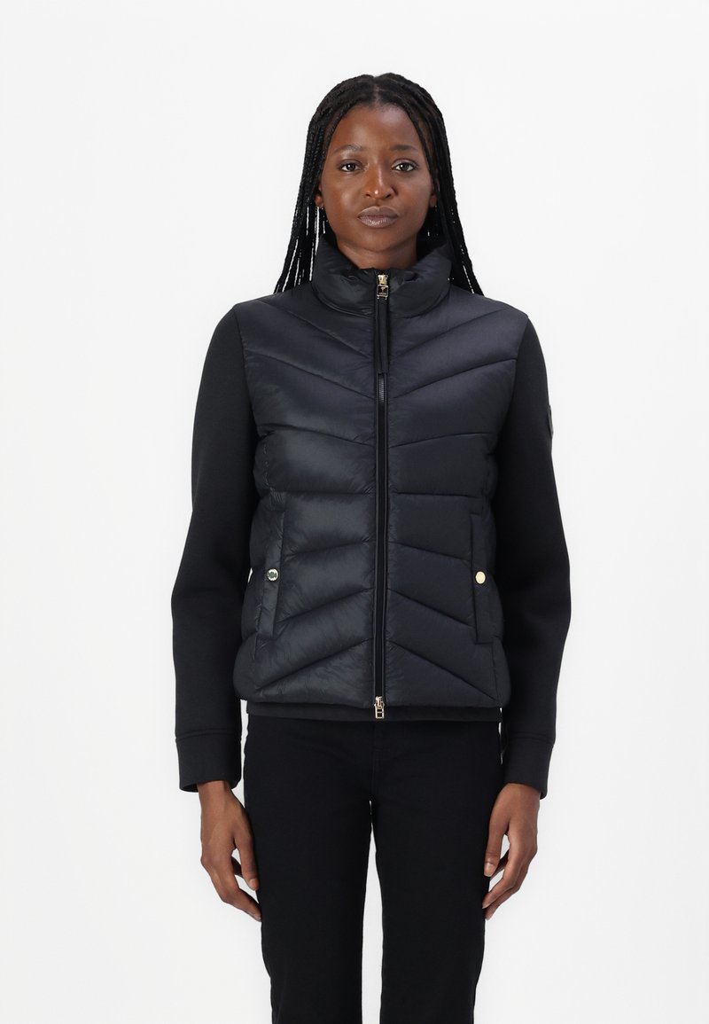 Vest puffer negru cu quilting în zigzag, guler înalt și închidere cu fermoar. Mâneci lungi dintr-un material neted. Accente de hardware aurie.