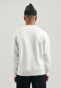 Felpa bianca con una vestibilità rilassata, polsini e orlo a coste, e un piccolo e discreto dettaglio di cucitura sul retro. Indossata con pantaloni scuri e un berretto.