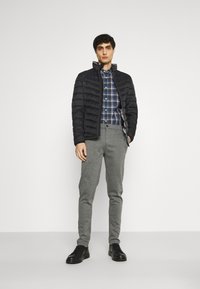 Doudoune noire sur une chemise à carreaux bleue et blanche, associée à un pantalon gris et des baskets noires. Texture lisse, design décontracté.