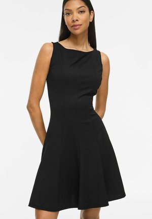 Femme aux longs cheveux foncés portant une robe noire sans manches, longueur genou, debout devant un fond blanc uni.