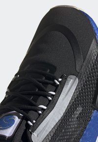 Svart träningsskor med en texturerad övre del, grå detaljer, snörning och blå inslag på sulan. Har en slät tådel.