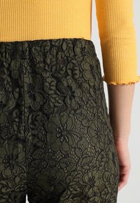 Czarne koronkowe spodnie w kwiatowy wzór, z elastycznym pasem i oliwkową podszewką widoczną przez koronkową teksturę.