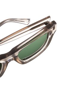 Occhiali da sole con montatura trasparente, lenti verdi e dettagli in metallo. Presentano una forma distintiva con curve pronunciate e cerniere eleganti.