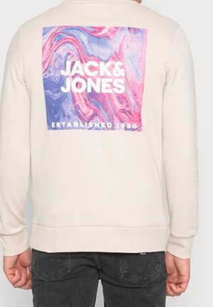 Beige sweatshirt med ett mönster av flerfärgad marmor på ryggen som har "JACK & JONES" och "ETABLERAD 1990" i fet vit text.