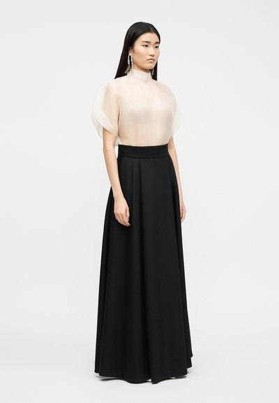 Femme portant un chemisier beige transparent à manches courtes avec un col montant et un pantalon noir large à jambes longues, devant un fond uni.