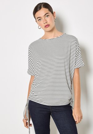 Mujer de cabello oscuro con una camiseta de manga corta a rayas blancas y negras con lazos en los lados, vistiendo jeans oscuros y pendientes de aro plateados, sobre un fondo blanco.