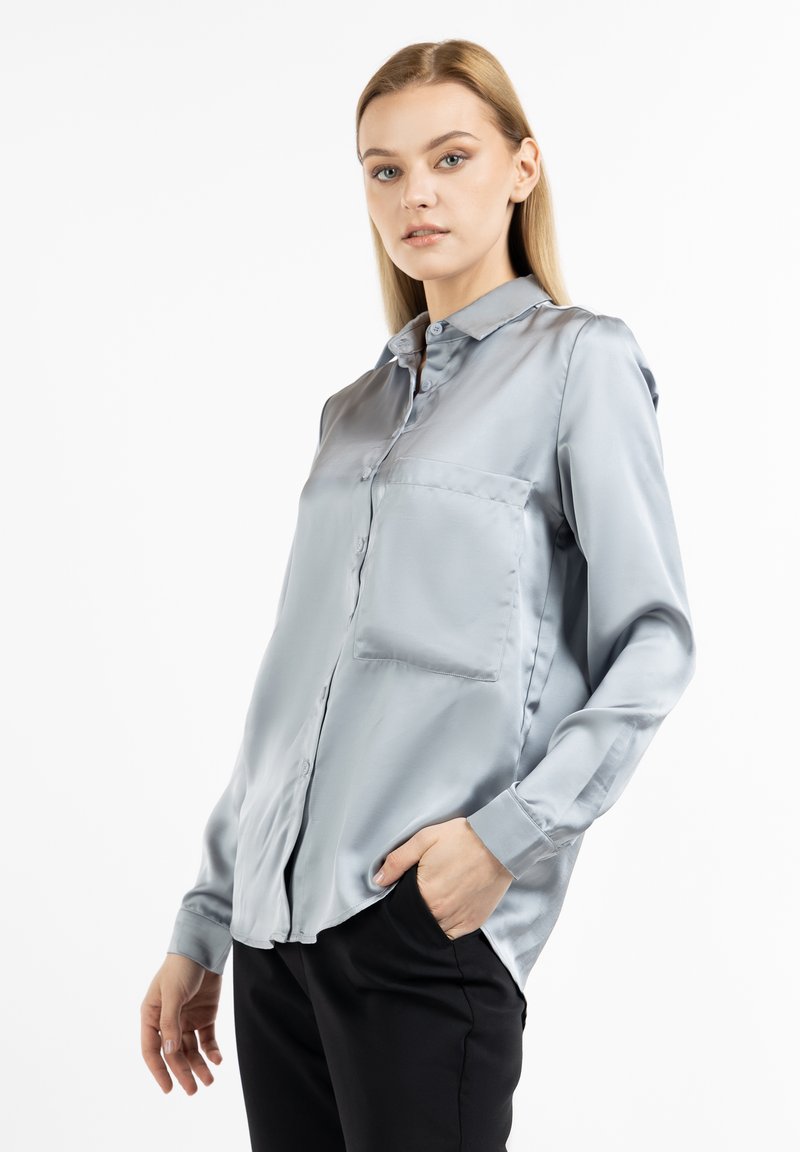 Blusa in satin azzurro chiaro con scollatura a colletto, chiusura con bottoni frontale e una tasca sul petto; maniche lunghe con polsini abbottonati.