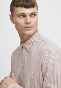 Cămașă cu dungi, cu nasturi, din bumbac de culoare maro deschis și alb. Prezintă un guler ascuțit și o textură subtilă, cu nasturi și detalii de cusătură vizibile.