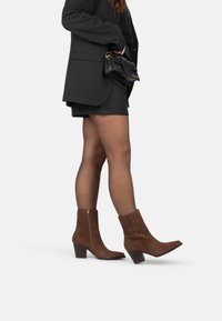 Botas de ante marrón hasta el tobillo con punta afilada, tacón ancho y cremallera lateral. Llevadas con pantalones cortos negros y un blazer, combinadas con un bolso negro.