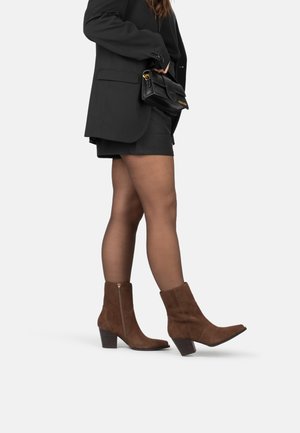 Botas de ante marrón hasta el tobillo con punta afilada, tacón ancho y cremallera lateral. Llevadas con pantalones cortos negros y un blazer, combinadas con un bolso negro.