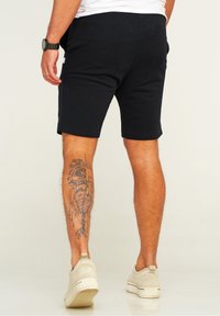 Black cotton shorts with a fitted design, visible pockets, and a small logo. Wearing beige suede slip-on shoes. Decorative tattoo on leg.