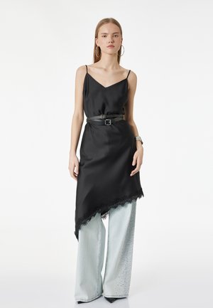 Jeune femme portant une robe nuisette noire asymétrique sur un jean large clair orné de strass, accessoirisée avec de grandes boucles d'oreilles anneaux et une double ceinture.