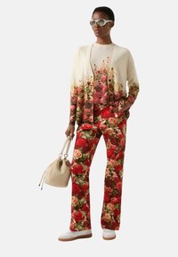 Kardigan dzianinowy w kwiatowy wzór z czerwonym i kremowym gradientem, zestawiony z dopasowanymi kwiatowymi spodniami. Akcesoria to beżowa torba i białe trampki.