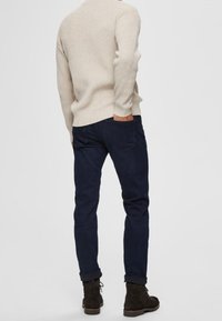 Světlý béžový žebrovaný svetr v kombinaci s tmavě modrými džínami a hnědými kotníkovými botami. Pohled zezadu, zobrazující horní část těla.