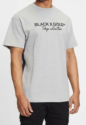 Mand iført en lysegrå T-shirt med teksten "BLACK X GOLD Tokyo collection" i sort, kombineret med sorte cargobukser.