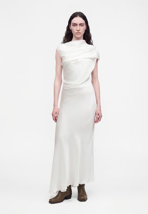 Femme aux longs cheveux foncés portant une robe blanche drapée sans manches et des bottines marron, debout devant un fond blanc uni.
