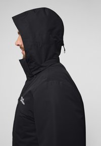 Veste noire à capuche avec fermeture éclair, conçue avec une texture lisse et une silhouette ajustée. Dispose d'un logo sur la poitrine et d'une capuche réglable.
