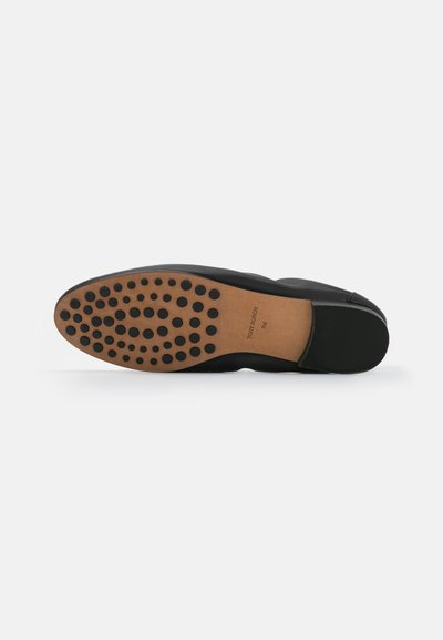 Chaussure noire avec un dessus lisse et une semelle extérieure en caoutchouc comportant des motifs de grip circulaires. La semelle intérieure est marron clair avec un logo.