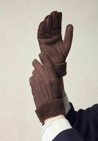 Brown suede gloves with a soft, plush cuff, featuring stitched detailing on the fingers. Durable material and structured design.