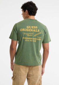 Grünes Baumwoll-T-Shirt mit gelbem Text und grafischen Elementen auf der Rückseite, einschließlich eines Schraubendesigns und Kontaktinformationen, runder Ausschnitt.