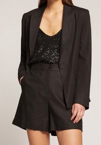 Černý lněný blejzr přes černý top s jemnými flitry a odpovídající lněné šortky. Tento outfit má precizně střižené linie a uvolněný střih.