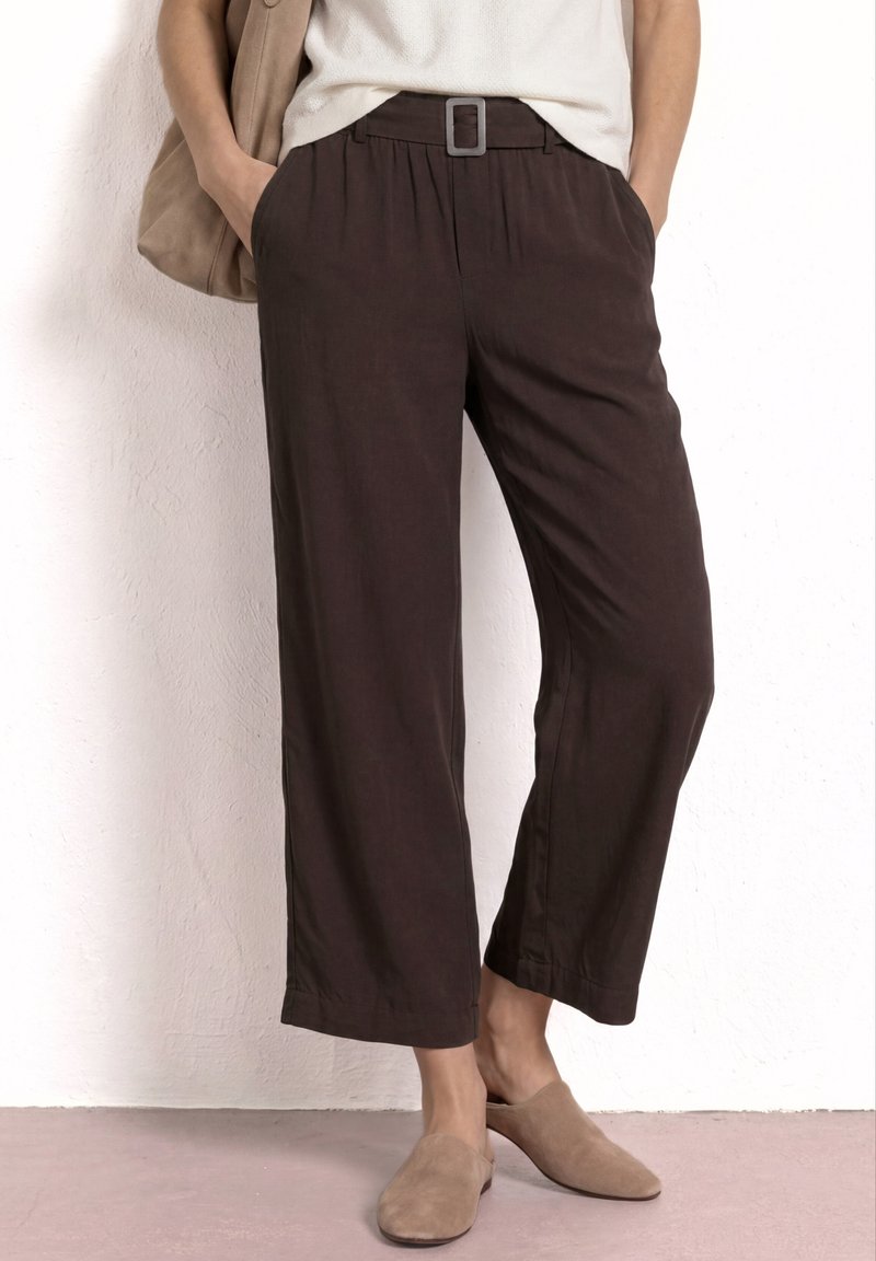 Vrouw draagt donkerbruine cropped rechte broek met riem, beige instapschoenen en een lichte top, staand tegen een witte muur.