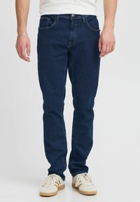 Mørkeblå jeans laget av denim, med rette ben, fem-lommers design og søm detaljer. Brukt med hvite joggesko.