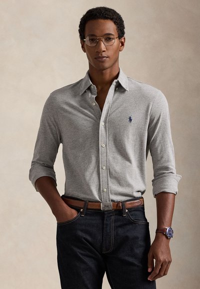Mand iført grå langærmet skjorte med knapper og opkrøllede ærmer, mørke jeans, brunt bælte, ur og runde briller foran en ensfarvet baggrund.