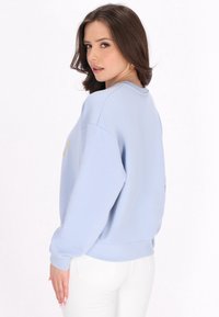Světle modrý svetr s kleslými rameny a zlatým nápisem, doplněný bílými slim-fit kalhotami. Měkká textura a uvolněný střih.