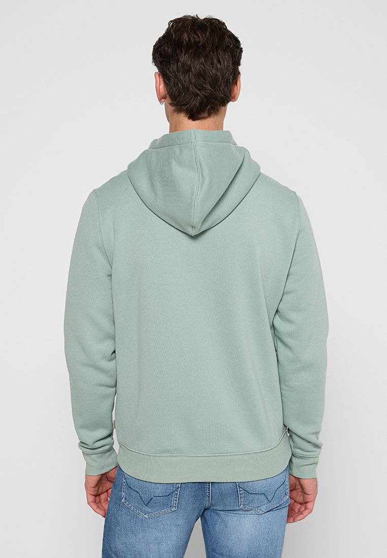 Sudadera verde menta con un tejido texturizado, puños y dobladillo acanalados, y capucha con cordón, combinada con unos jeans de mezclilla azul. Vista trasera mostrada.