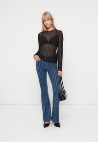 Zwarte polka dot mesh top met lange mouwen, gecombineerd met blauwe streepjasje flared jeans. Model houdt een kleine zwarte handtas vast en draagt zwarte hakken.
