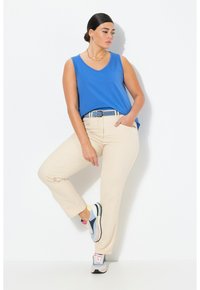 Blauw mouwloos topje, beige broek met een riem. Sneakers met blauwe, gele en roze accenten. Model staat met één voet naar voren tegen een witte achtergrond.