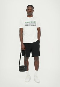 Balta trumpa rankovėmis marškinėliai su daugiakomponiu "ARMANI EXCHANGE" logotipu, juodos šortai, juoda per petį nešama kuprinė ir balti sportbačiai su tekstūruotais padais.